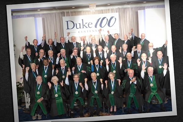 Portrait of elder alumni in green regalia with a Duke 100 centennial banner in the background